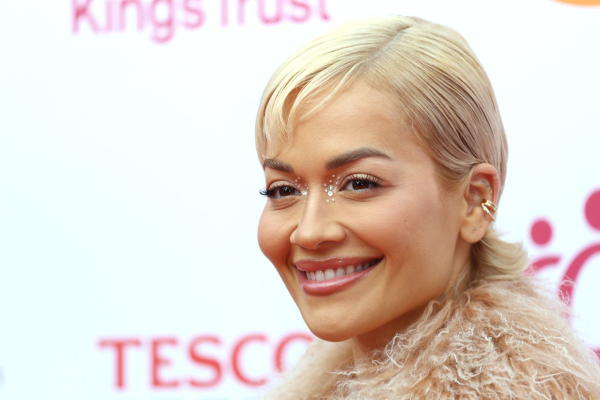 Rita Ora wears eye gems in her inner corners. She has blonde hair and wears a furry top.
